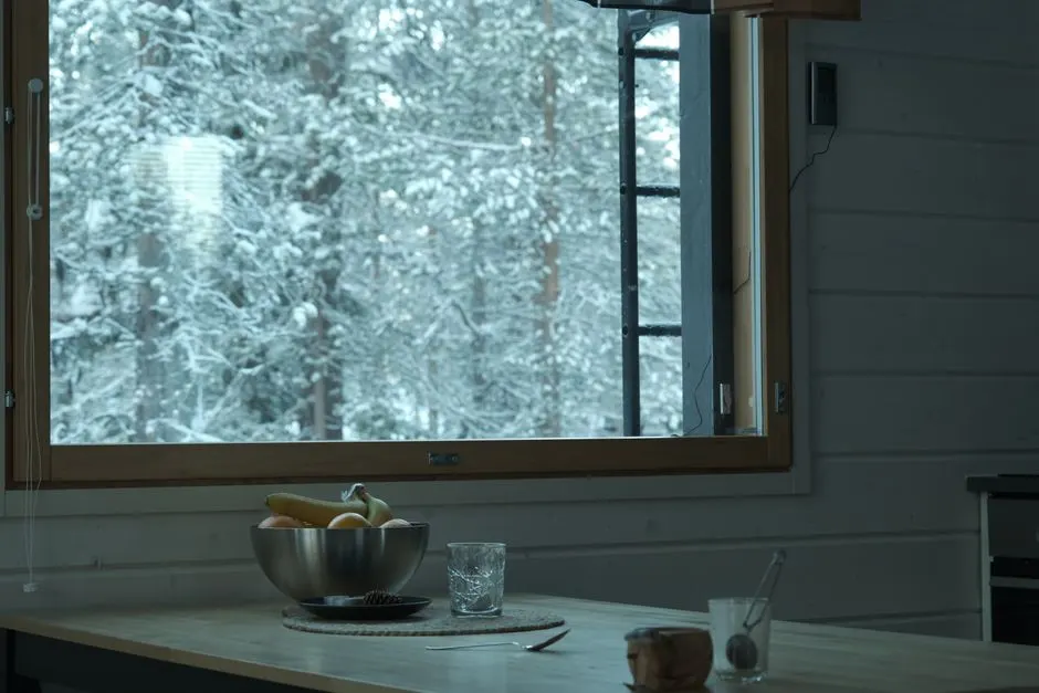 A serene kitchen with a snowy forest view through a large window, featuring a bowl of fruit on the table.